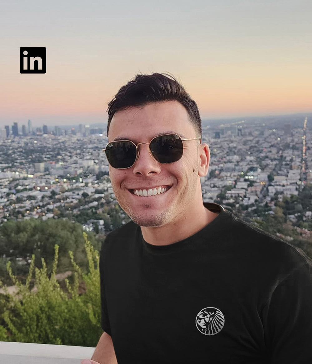 Anthony, co-founder of Tupan, smiling in front of the Los Angeles skyline while wearing a black shirt with the tribal Tupan crest — one of the warriors behind açaí coffee.