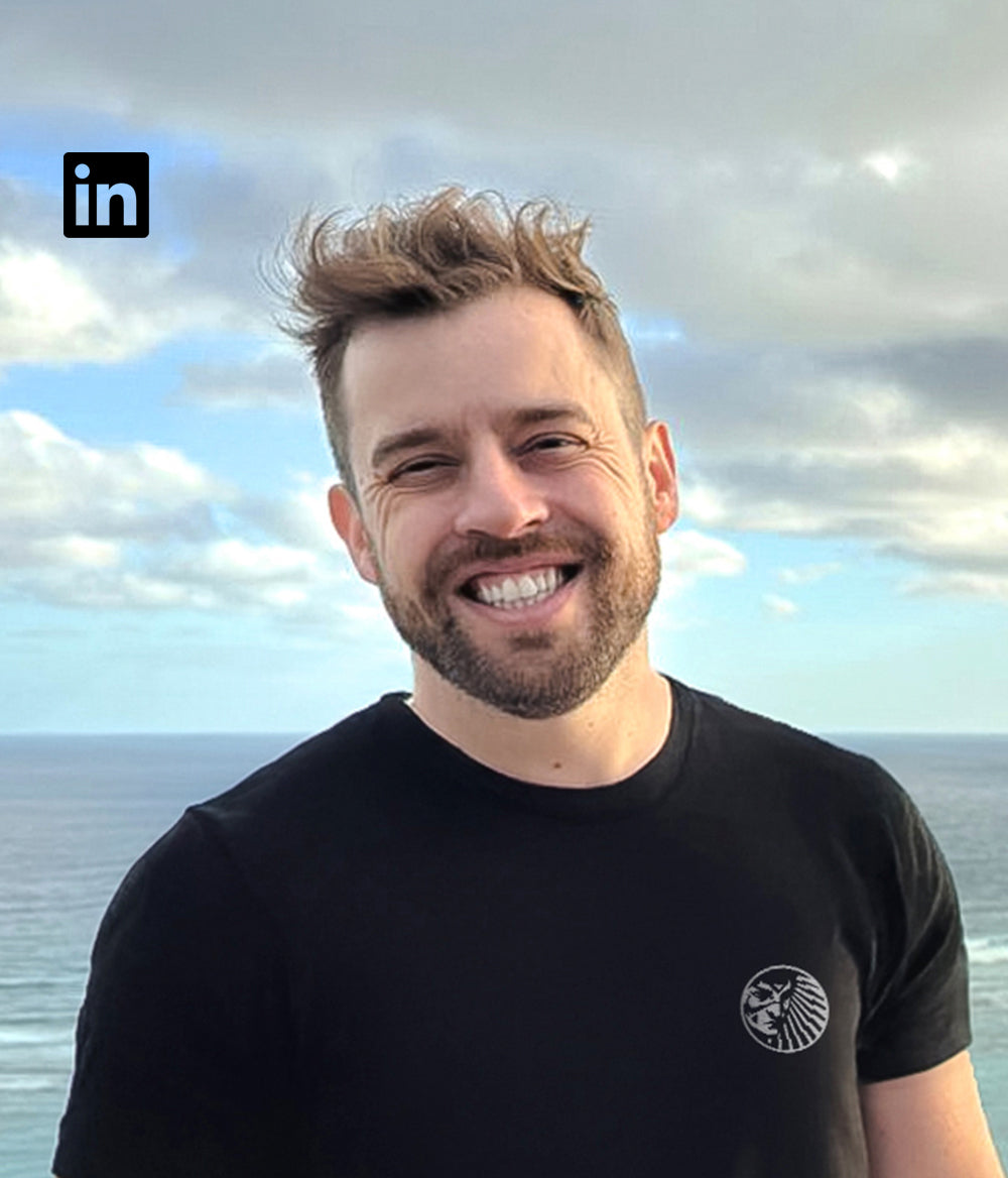Matheus, co-founder of Tupan, in front of the ocean with a wide smile, wearing a black shirt with the Tupan tribal symbol — a core force in the açaí coffee tribe.